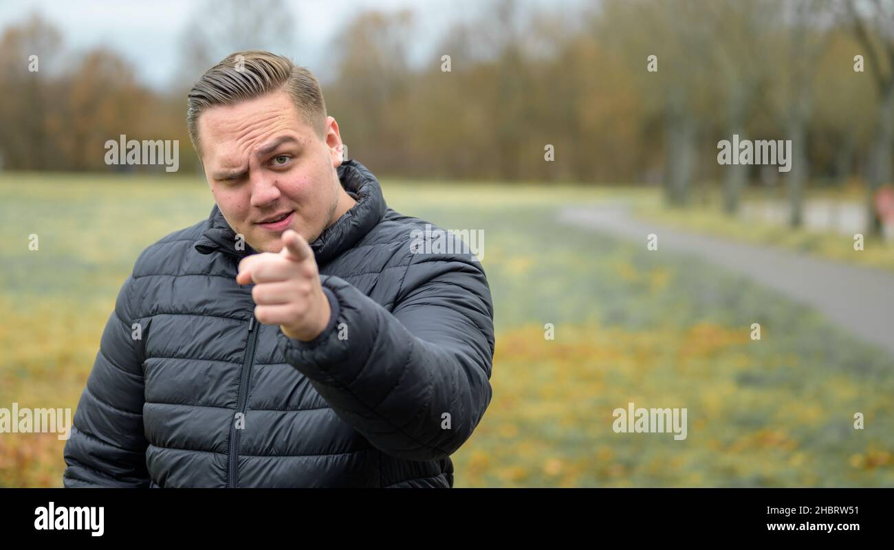 Ein junger Mann, der im Herbst in einem Park mit einem Finger auf die Kamera zeigt, zeigt Skepsis und ein zweifelhaftes Stirnrunzeln Stockfoto