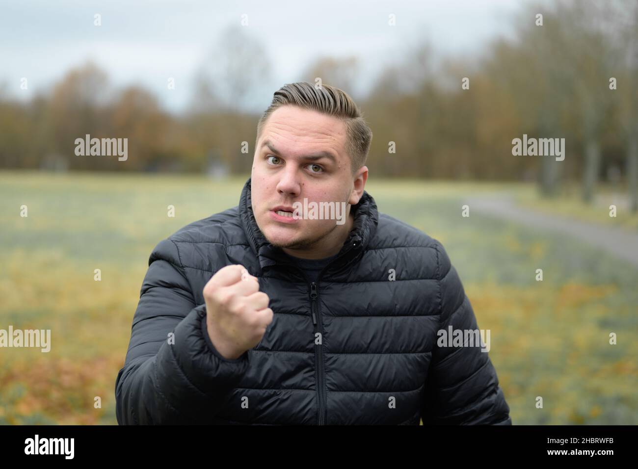 Wütender junger Mann, der die Kamera mit der Faust bedroht, während er im Freien in einem herbstlichen Park mit einem balsamiger Blendung schaust Stockfoto