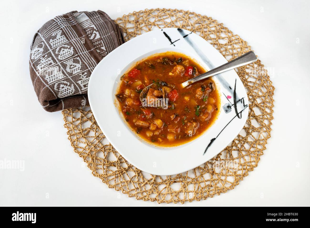 Reichhaltige und würzige ländliche rote Suppe mit Schweinefleisch, rotem Pfeffer, Tomaten, Pilzen, Gemüse und Bohnen auf dem Teller, Handtuch und Bambuskissen auf weißem Hintergrund. Stockfoto