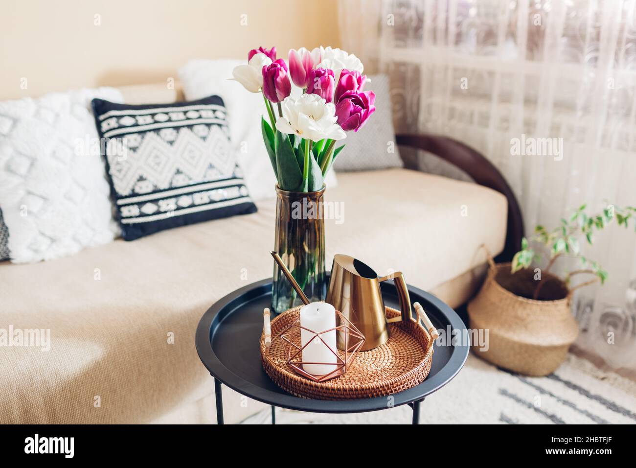 Inneneinrichtung und Einrichtung im Frühling. Frische Tulpen Blumen setzen in Vasen auf Couchtisch mit Kerze und Gießkanne auf Rattan Tablett. Gemütliches Wohnzimmer Stockfoto
