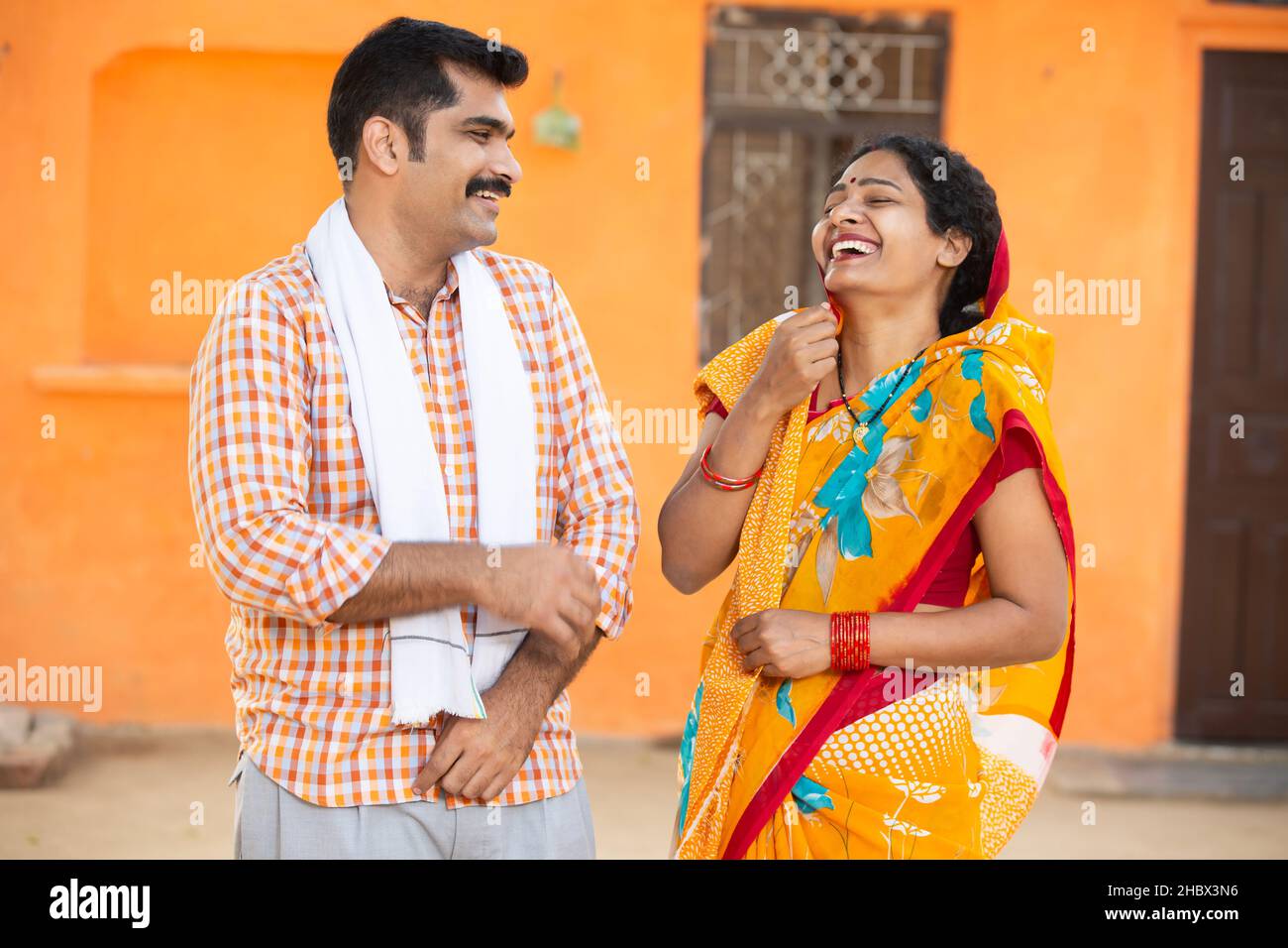 Junges indisches Landpaar, das vor dem Dorfhaus lachend steht. Schnurrbart, Mann mit Kurta und Frau mit Sari, Ehemann, Frau mit Traditio Stockfoto