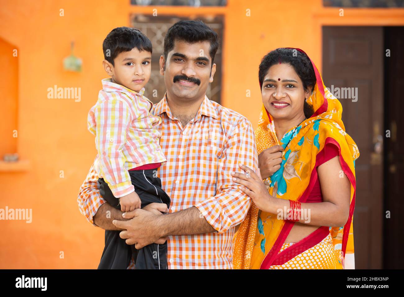 Porträt einer jungen indischen Landfamilie, die im Dorfhaus im Freien steht. Eltern mit ihrem kleinen Kind, Schnurrbart-Mann in Kurta und Frau tragen Stockfoto