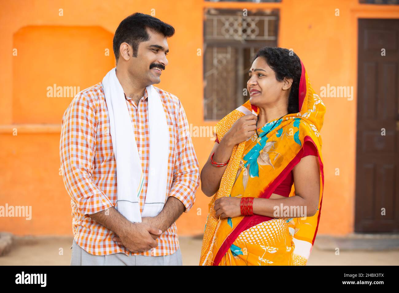 Junges indisches Landpaar, das vor dem Dorfhaus lachend steht. Schnurrbart, Mann mit Kurta und Frau mit Sari, Ehemann, Frau mit Traditio Stockfoto