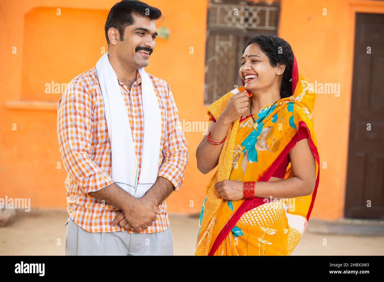 Junges indisches Landpaar, das vor dem Dorfhaus lachend steht. Schnurrbart, Mann mit Kurta und Frau mit Sari, Ehemann, Frau mit Traditio Stockfoto