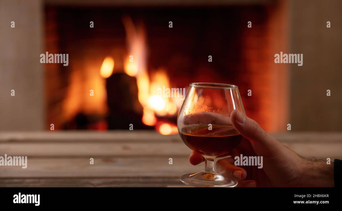 Trinken Sie Alkohol und entspannen Sie sich im Winter. Mann mit einem Brandy Glas, Holztisch und brennendem Kamin Hintergrund, Platz. Warme, gemütliche Inneneinrichtung Stockfoto