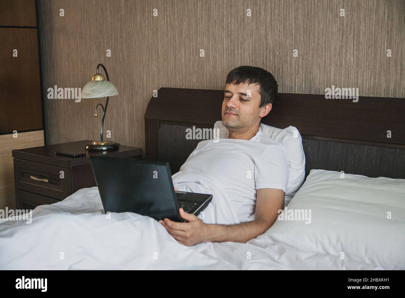 Ein Mann mittleren Alters sitzt im Bett in seinem Schlafzimmer und schaut sich ein Video auf einem Laptop an. Stockfoto