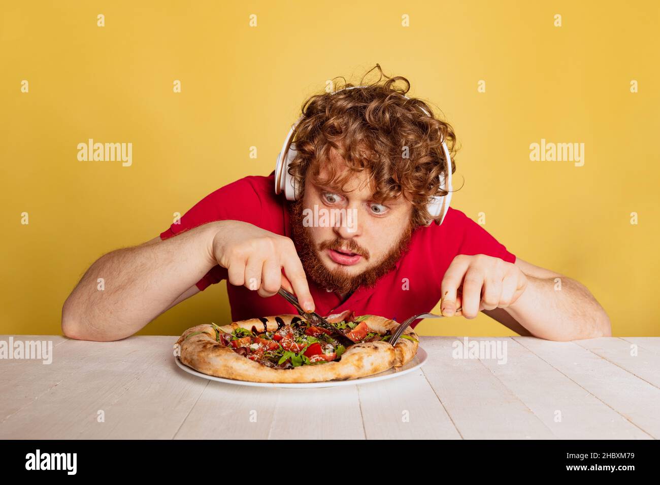 Schockierter rotbärtiger Mann schneidet und isst leckere Pizza mit Käse und Tomaten isoliert auf gelbem Studiohintergrund. Stockfoto