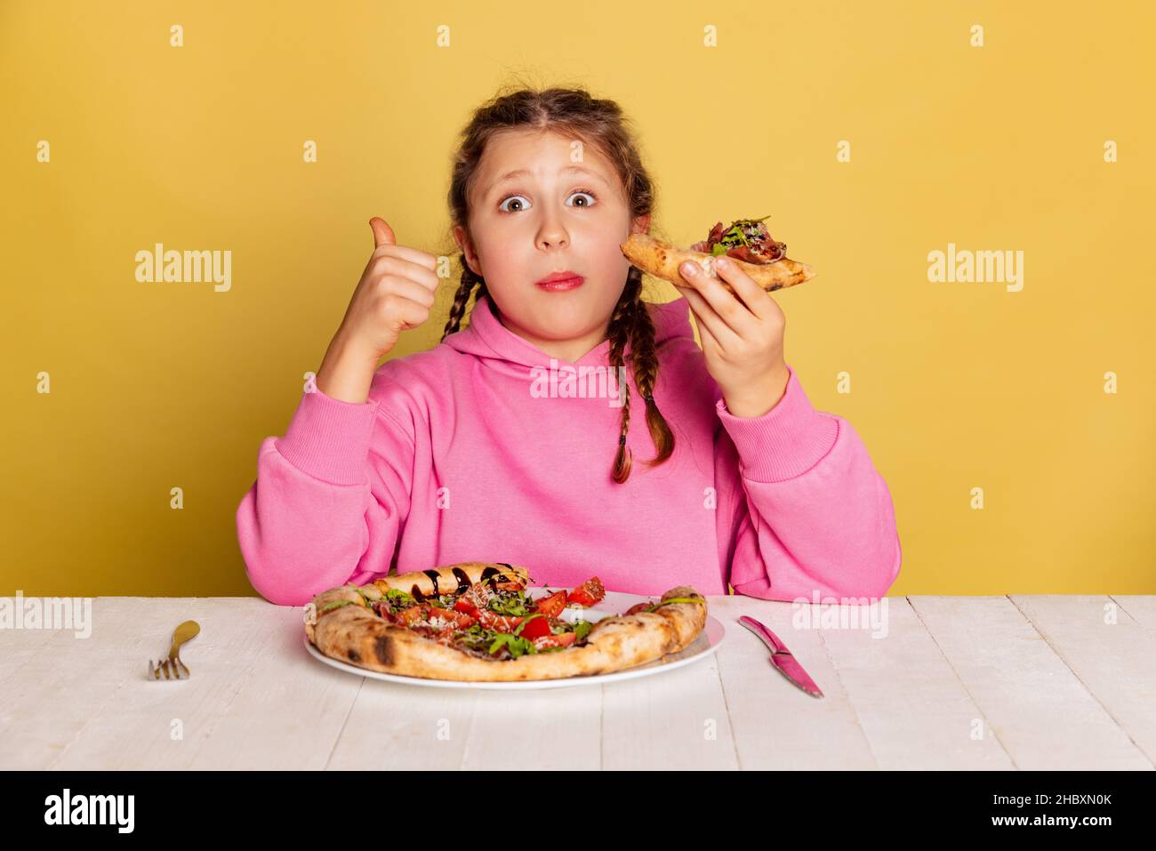 Studio-Aufnahme von kleinen niedlichen Mädchen sitzen und Verkostung köstliche italienische Pizza isoliert auf gelben Studio-Hintergrund. Stockfoto