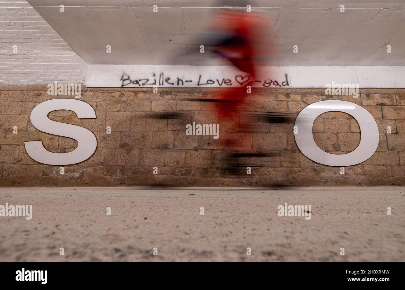 22. Dezember 2021, Sachsen, Chemnitz: Eine Frau fährt mit dem Fahrrad durch die neu gestaltete Bazillenröhre, den Fußgängertunnel am Chemnitzer Hauptbahnhof. Der enge Tunnel zwischen der Innenstadt und dem Stadtteil Sonnenberg wird nun unter anderem mit einem neuen Lichtkonzept aufgewertet. Zudem bilden 1,40 Meter hohe weiße Buchstaben an den Wänden ein Zitat aus dem Song 'wie ich' der Band Kraftklub: 'Ich wär'gern weniger wie ich, ein bisschen mehr so wie du'. Im Volksmund wird die früher eher dunkle, 220 Meter lange Unterführung im Volksmund als „Keimröhre“ bezeichnet. In zwei Konstr Stockfoto