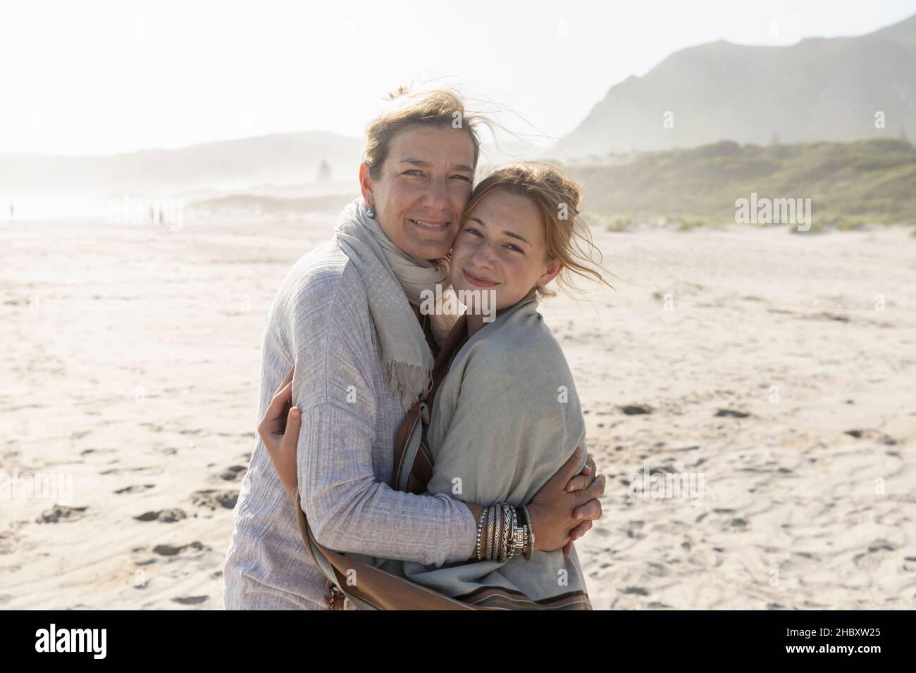 Die Erwachsene Frau und ihre Tochter im Teenageralter umarmen sich und stehen an einem windgepeitschten Strand Stockfoto