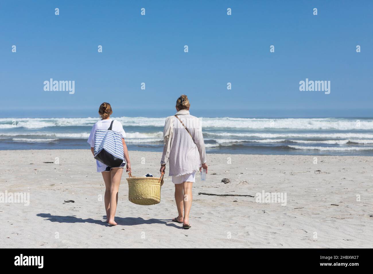 Mutter und Tochter im Teenageralter, die mit einem Korb an einem Sandstrand spazieren gehen Stockfoto