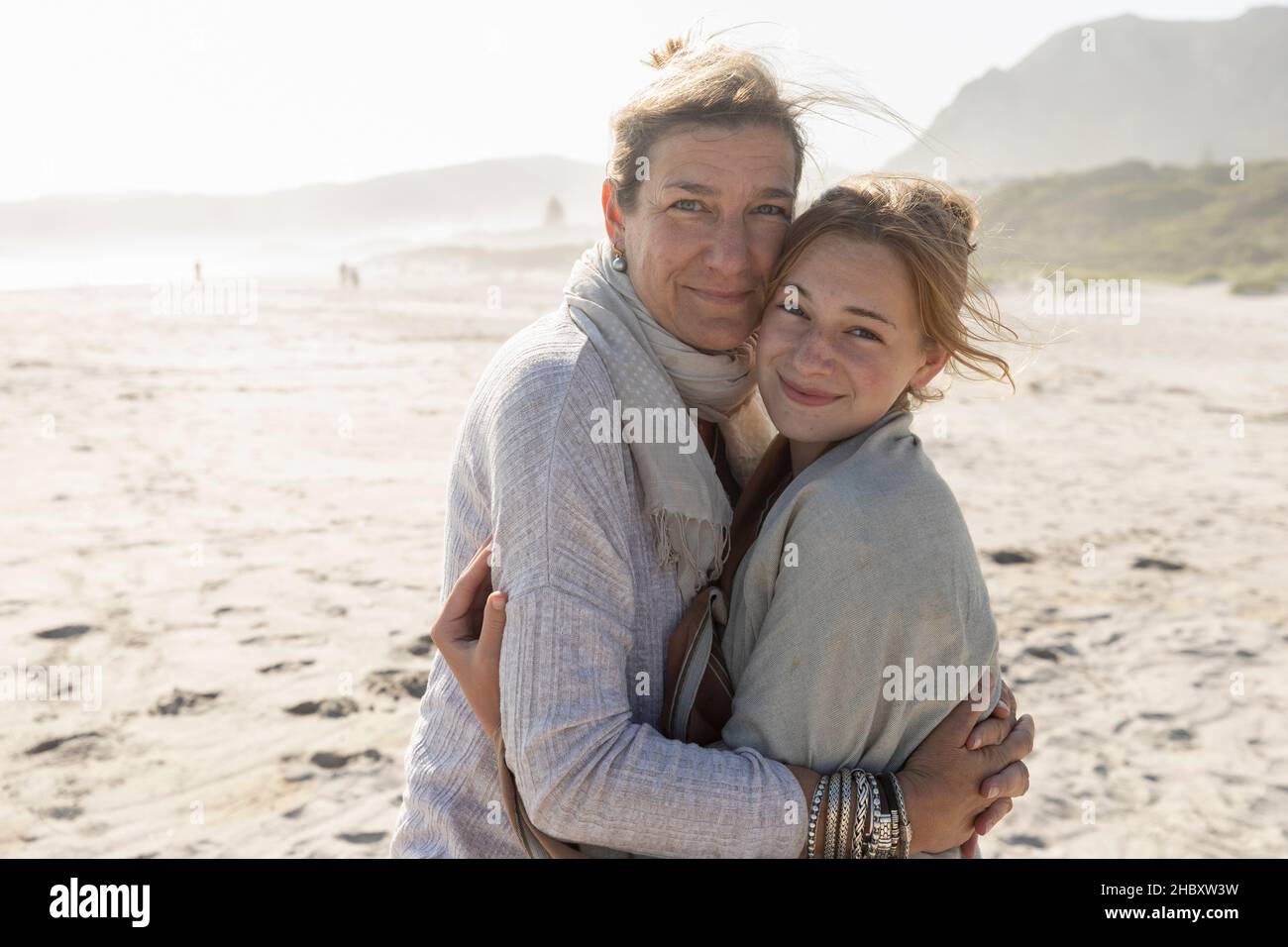 Die Erwachsene Frau und ihre Tochter im Teenageralter umarmen sich und stehen an einem windgepeitschten Strand Stockfoto
