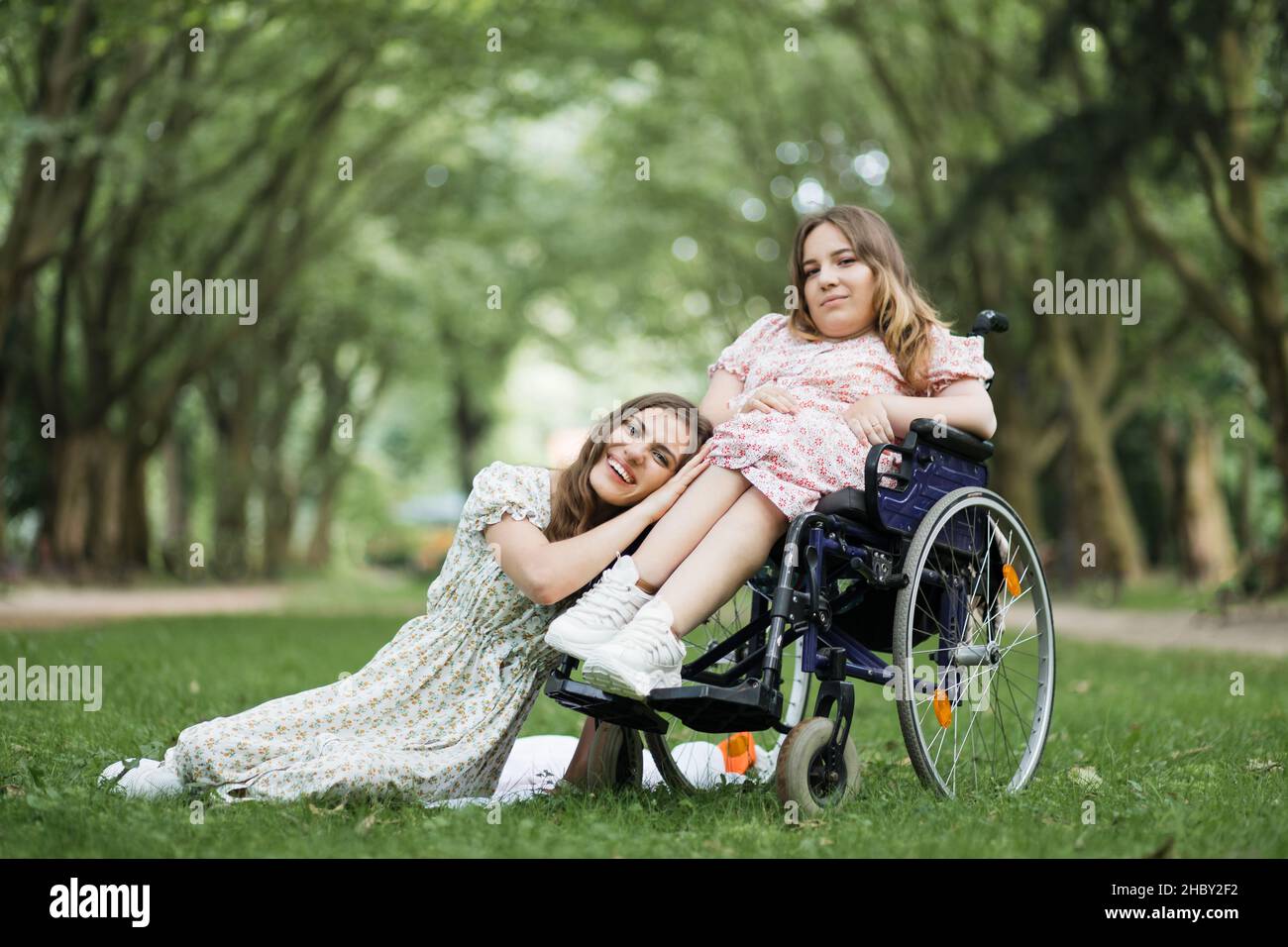 Hübsche kaukasische Frau, die auf Gras sitzt und den Kopf auf den Knien einer Freundin mit körperlicher Behinderung lehnt. Junge Dame im Rollstuhl fühlt sich sehr unterstützt und verständnisvoll. Stockfoto