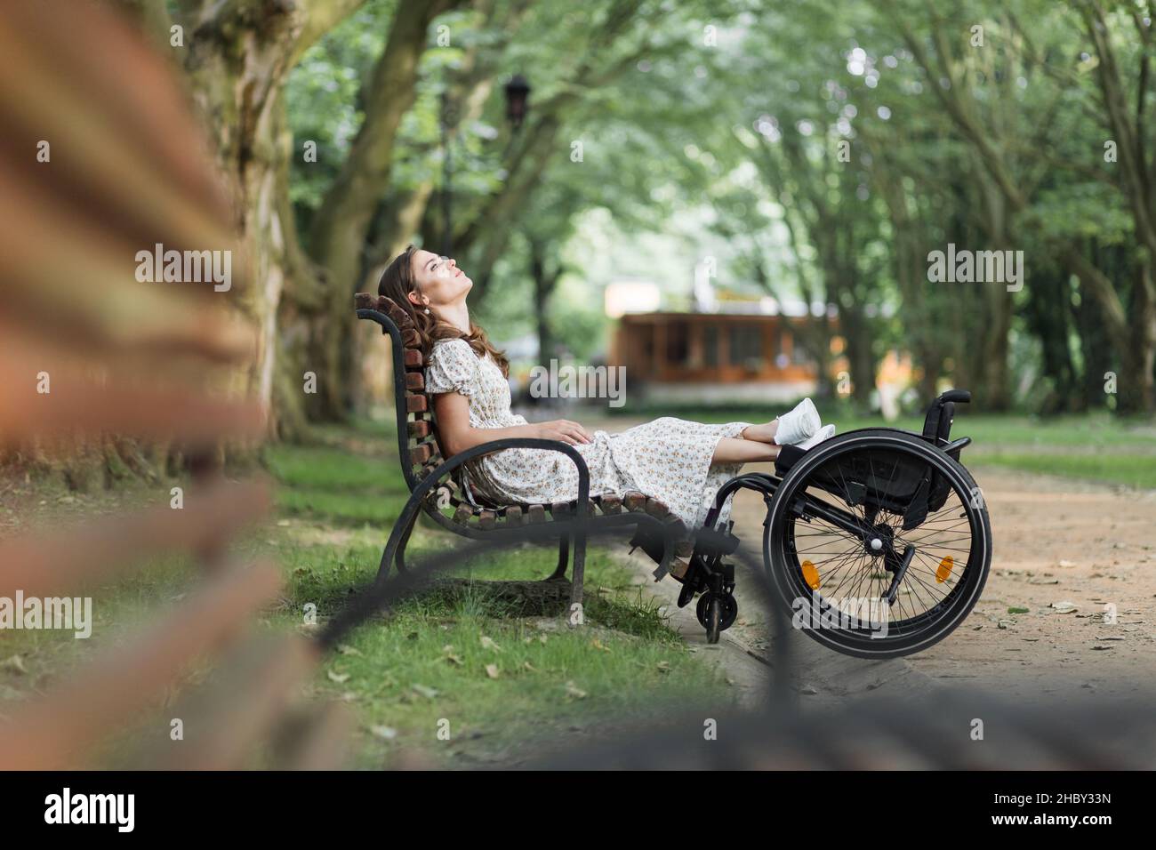 Kaukasische behinderte Frau, die sich auf einer Holzbank im Freien mit Füßen auf einem Rollstuhl ausruht. Hübsche junge Frau, die aufschaut und jeden Moment ihres Lebens genießt. Stockfoto