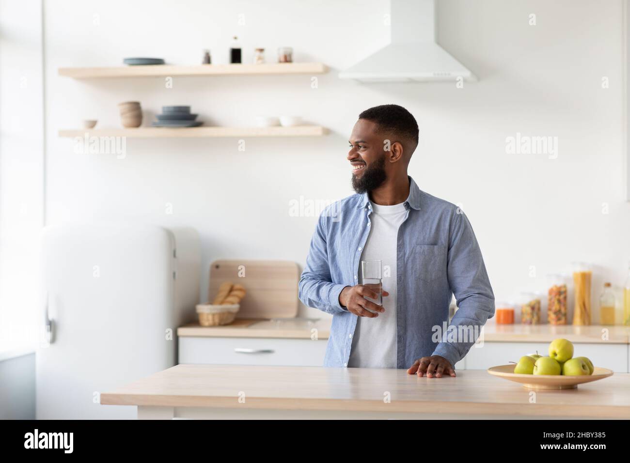 Fröhlicher junger bärtiger schwarzer Kerl mit einem Glas sauberem Wasser in der Hand in der Kücheninneneinrichtung schaut auf das Fenster Stockfoto