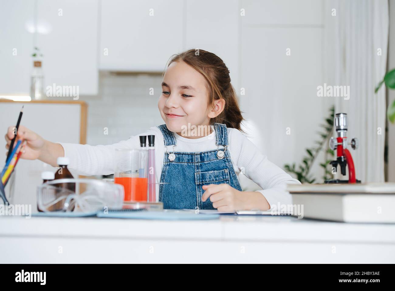 Lächelndes kleines Mädchen macht zu Hause Wissenschaft Projekt, nach Buntstiften zu greifen. Sie hat Chemiegläser mit bunten Flüssigkeiten und Mikroskop Stockfoto