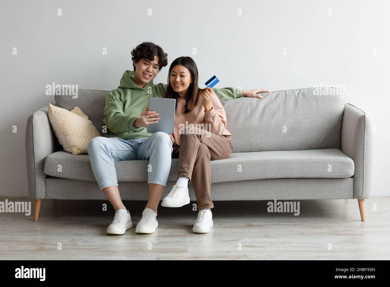 Junges asiatisches Paar mit Kreditkarte und Tablet auf dem Sofa sitzend, Waren im Internet bestellend, Sachen im Webshop kaufend Stockfoto