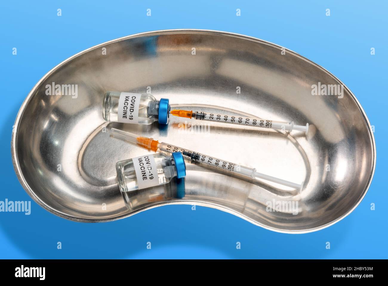 Zwei Impfstofffläschchen mit Spritze, die in einer nichtrostenden medizinischen Nierenschale ansaugt, auf hellblauem Hintergrund Stockfoto