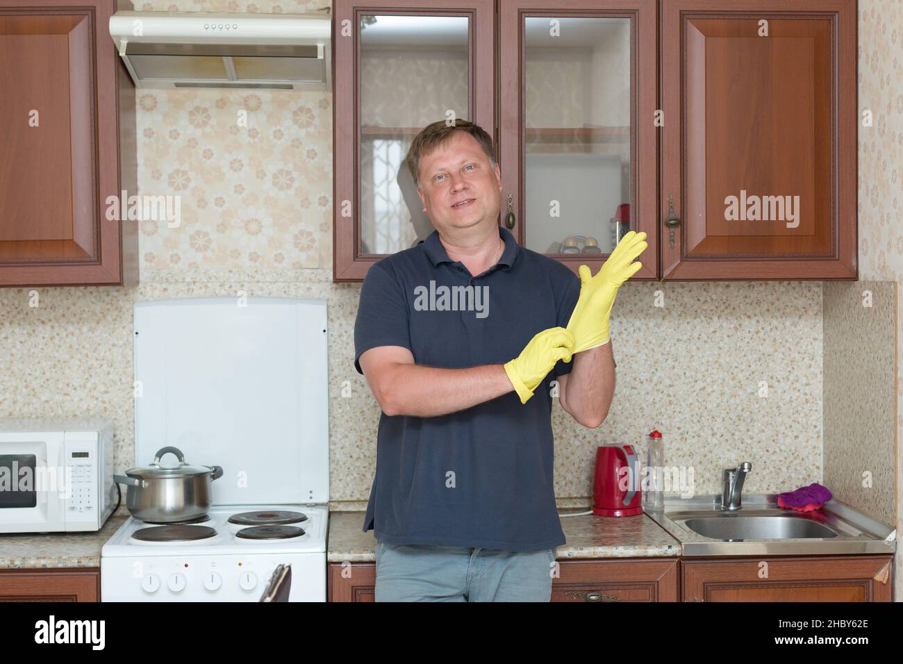 Ein großer erwachsener Mann mit gelben Gummihandschuhen in Hauskleidung putzt in der Küche. Stockfoto