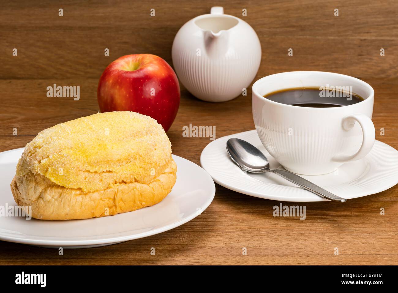 Frisches Butterbrot in weißer Keramikplatte, eine Tasse heißen schwarzen Kaffee mit Metalllöffel in Keramikuntertasse, einen reifen roten Apfel und einen weißen Milchtopf auf einem Holz Stockfoto