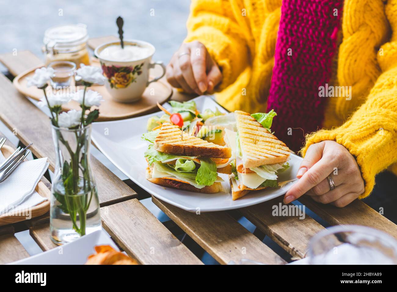 Frau, die im Café im Freien an der City Street frühstückte. Sandwich und Kaffee werden auf dem Tisch serviert. Frau in Strickpullover beim morgendlichen Restaurant Stockfoto