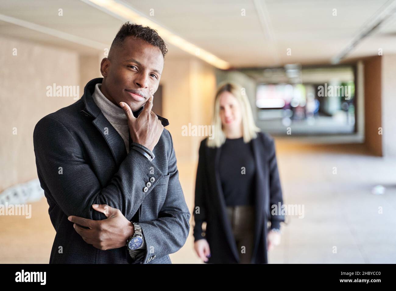 Afroamerikanischer Mann in stilvollem Mantel, der die Kamera anschaut, während er in einer modernen Passage in der Nähe einer Freundin steht Stockfoto
