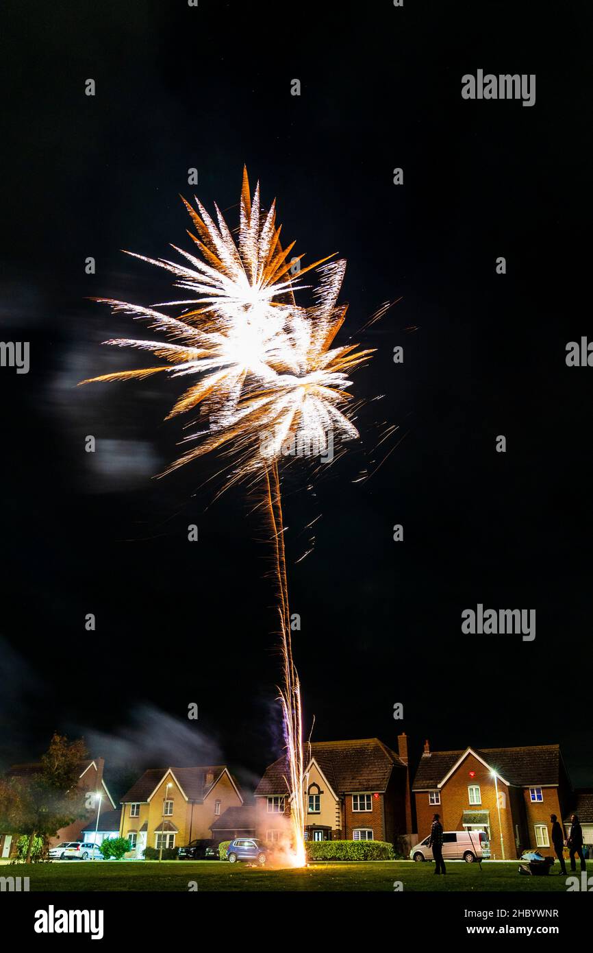 Feuerwerk auf Dorfgrün mit Feuerwerk explodiert auf einer goldenen feurigen Spur in abstraktem, verschwommenem Muster am Himmel über einer Reihe von Häusern. Stockfoto