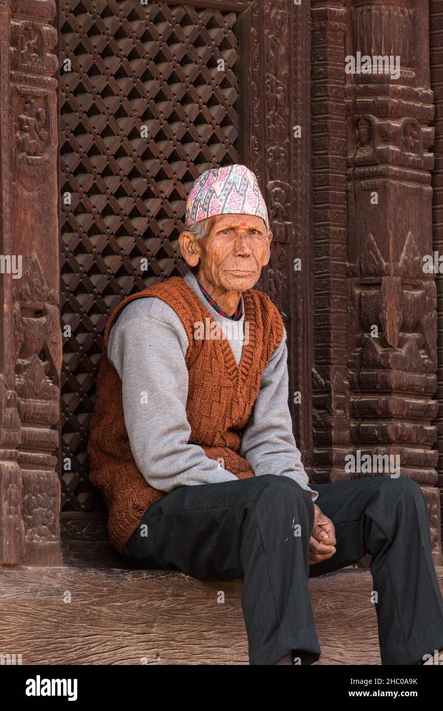 In der mittelalterlichen Stadt Bhaktapur in Nepal sitzt ein älterer Nepalesischer Mann mit einer Dhaka-Topi-Mütze vor kunstvoll geschnitzten Holzarbeiten. Stockfoto