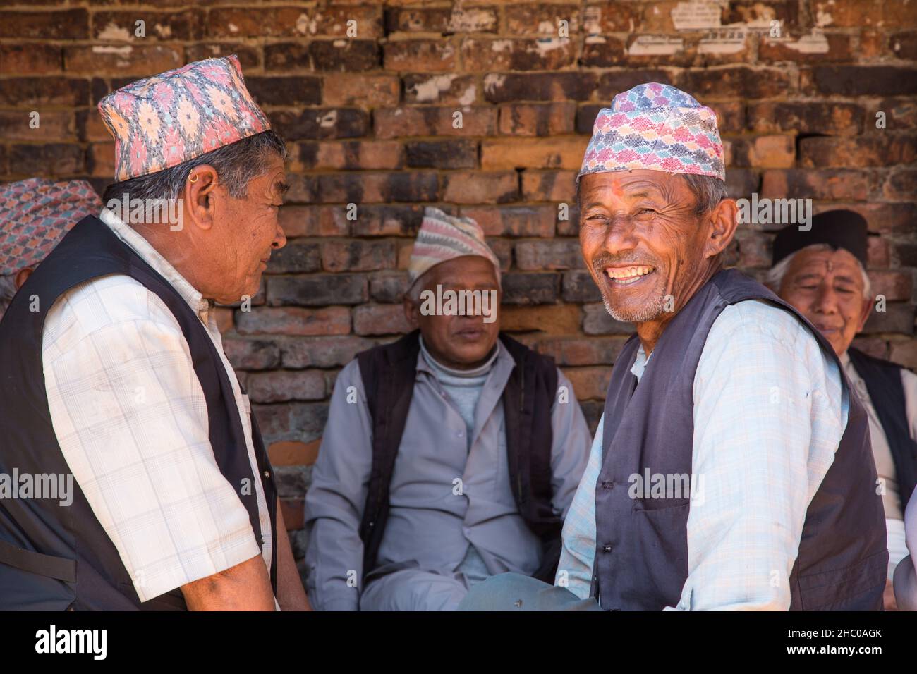 Eine Gruppe nepalesischer Männer in den traditonalen Dhaka-Topi-Kappen ruhen im Schatten und besuchen die mittelalterliche Stadt Bhaktapur in Nepal. Stockfoto