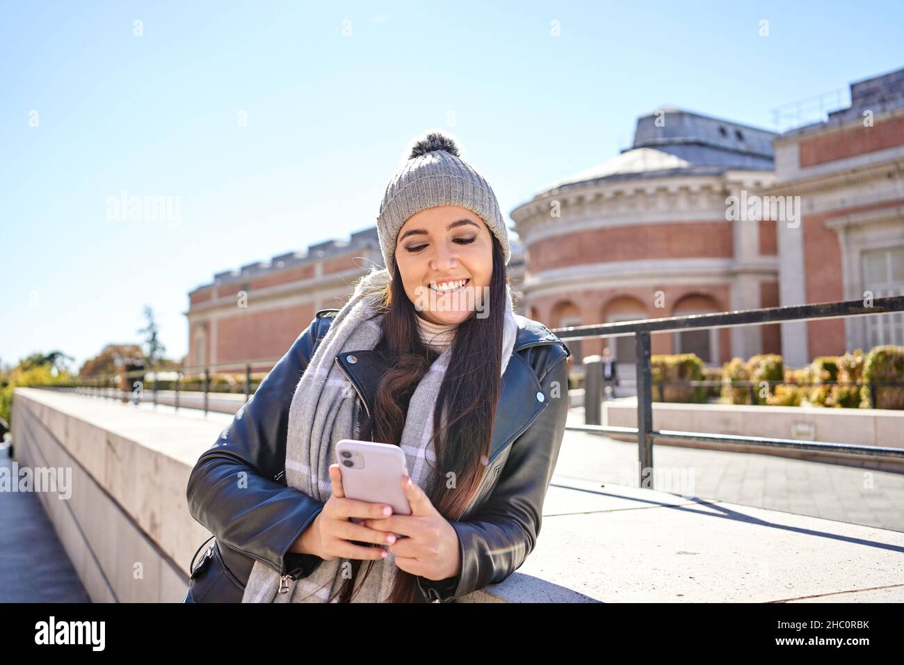 Selbstbewusste junge Frau, tausendjährig, mit langen dunklen Haaren in warmer Strickmütze und Lederjacke, lächelnd, während sie die Nachricht auf dem Mobiltelefon liest, das auf der Straße der Stadt vor wolkenlosem, blauem Himmel steht Stockfoto