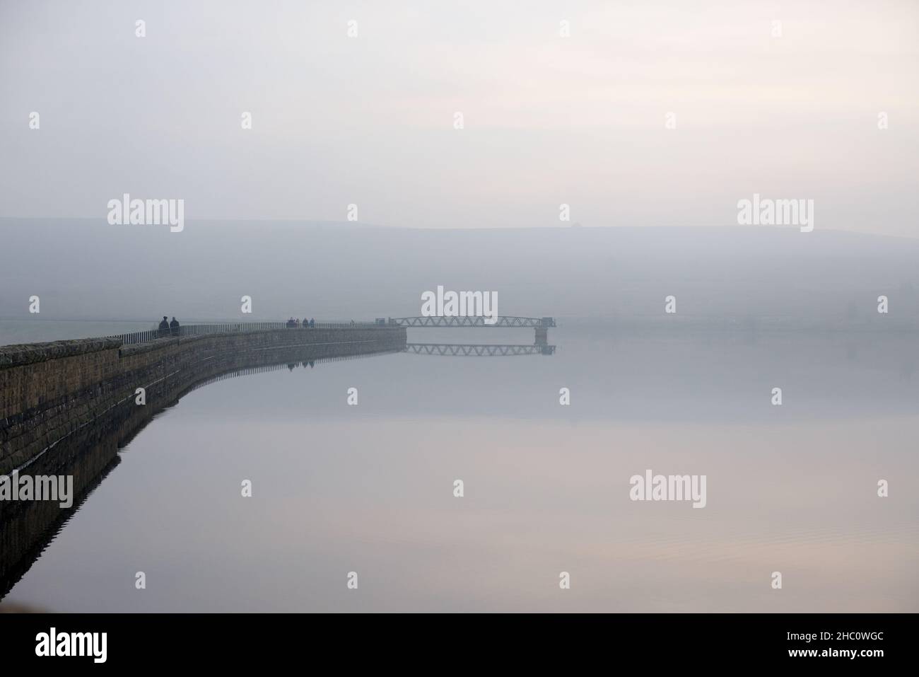 Sheffield, Großbritannien, 7th. November 2020: Düstere Winterszene über dem Rotmires-Reservoir. Gruppen von Menschen gehen an der Staumauer. atmosphärischer Nebel oder Nebel Stockfoto