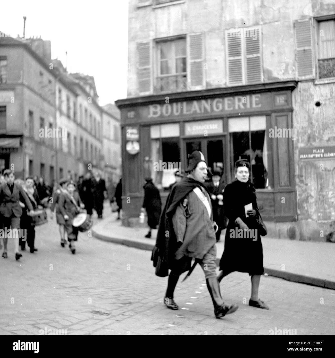 Eine Prozession im Jahr 1945 in Paris Montmartre kommt aus der Richtung Sacre-Cœur zu Fuß die Rue Norvins hinunter und links den Place Jean-Baptiste Clement hinunter. Führend ist ein älterer Mann, der einen Napoleonhut, einen Mantel und Reitstiefel trägt. Zigarre im Mund, ein Schwert baumelt an seiner linken Seite. Er scheint seinen rechten Arm verloren zu haben. Hinter ihm im marsch schlagen eine Gruppe von Jungen Trommeln. Ein von der US Navy eingesandter Mann beobachtet von einer Seite und Eine Frau beobachtet und schließt sich dann an die Spitze der Gruppe an. Der Konsulat d’Auvergne und das Restaurant L’ambassade und die Cafés Gilbert sind zu sehen Stockfoto