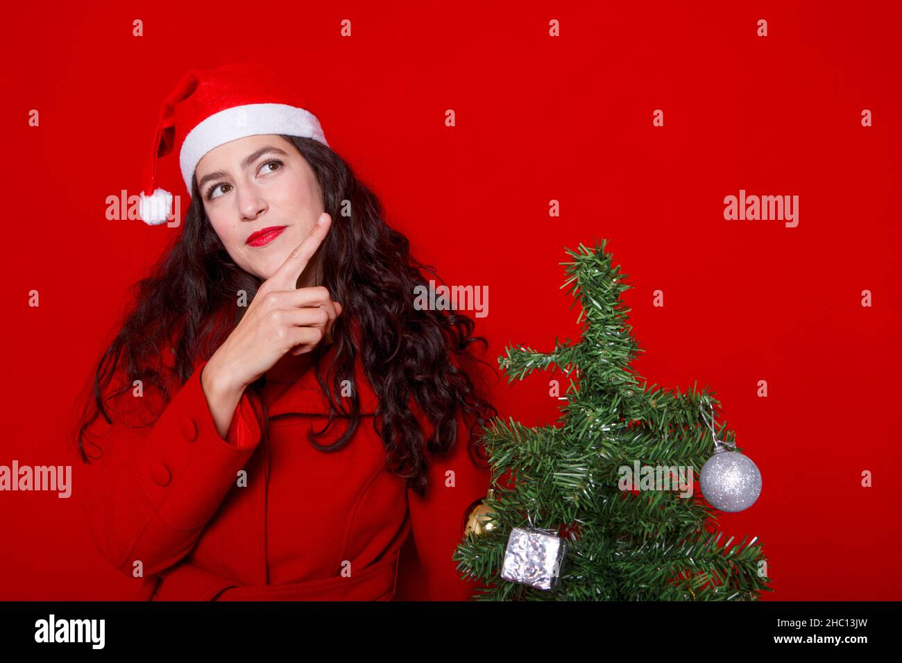 Lächelnde 37-jährige Frau im Mantel und Weihnachtshut zeigt die Finger, um den Raum isoliert auf rotem Hintergrund zu imitieren, Studioportrait. Frohes Neues Jahr Stockfoto