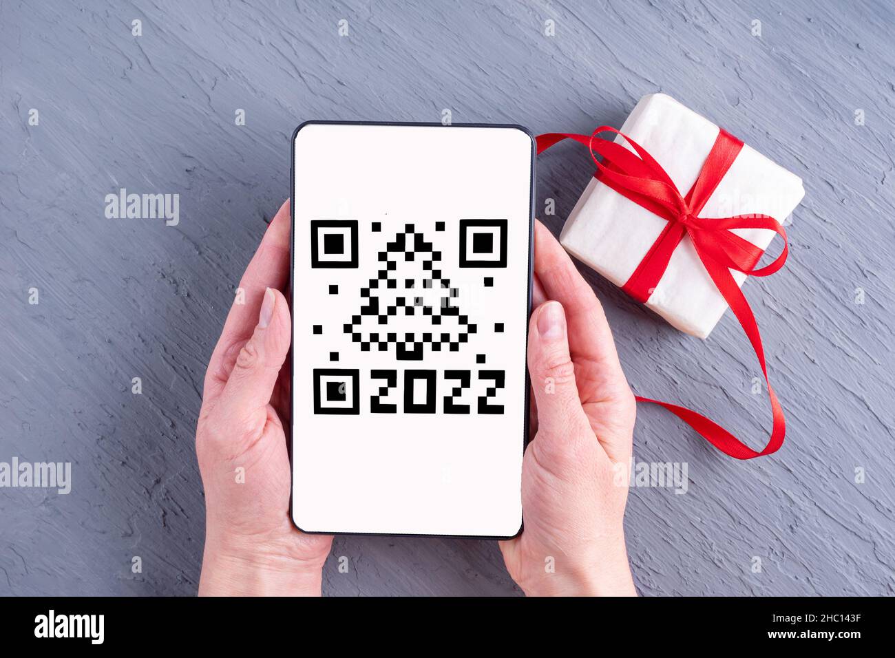 Hände, die ein Smartphone, ein Tablet, ein Mobiltelefon mit einem qr-Code-Bildschirm in Form eines Weihnachtsbaumdatums 2022 und einer Geschenkbox auf grauem Hintergrund halten. Stockfoto