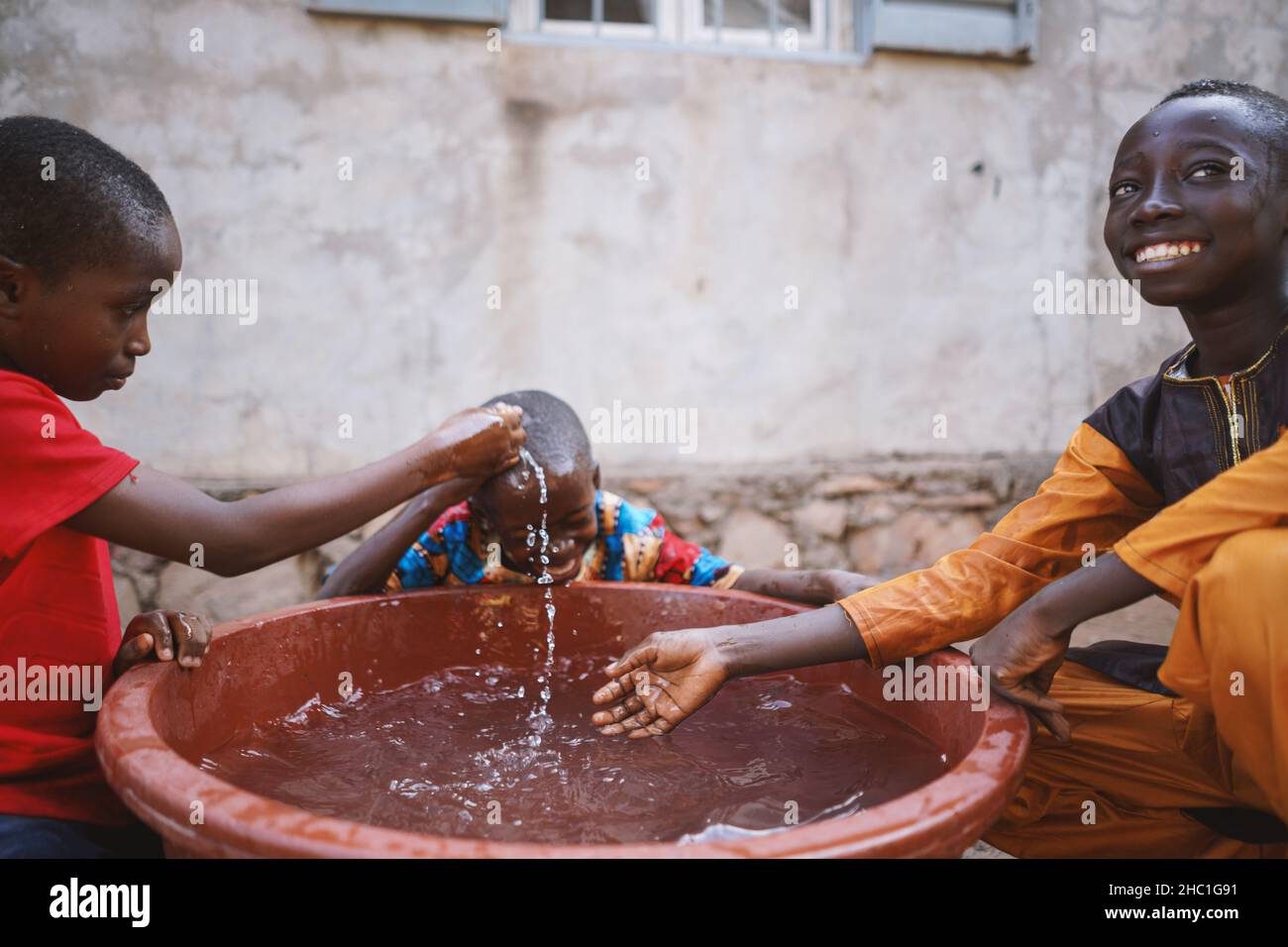 Drei kleine schwarze Jungen, die mit Wasser spielen, sitzen irgendwo in Westafrika um eine große braune Wanne vor dem Haus ihrer Eltern Stockfoto
