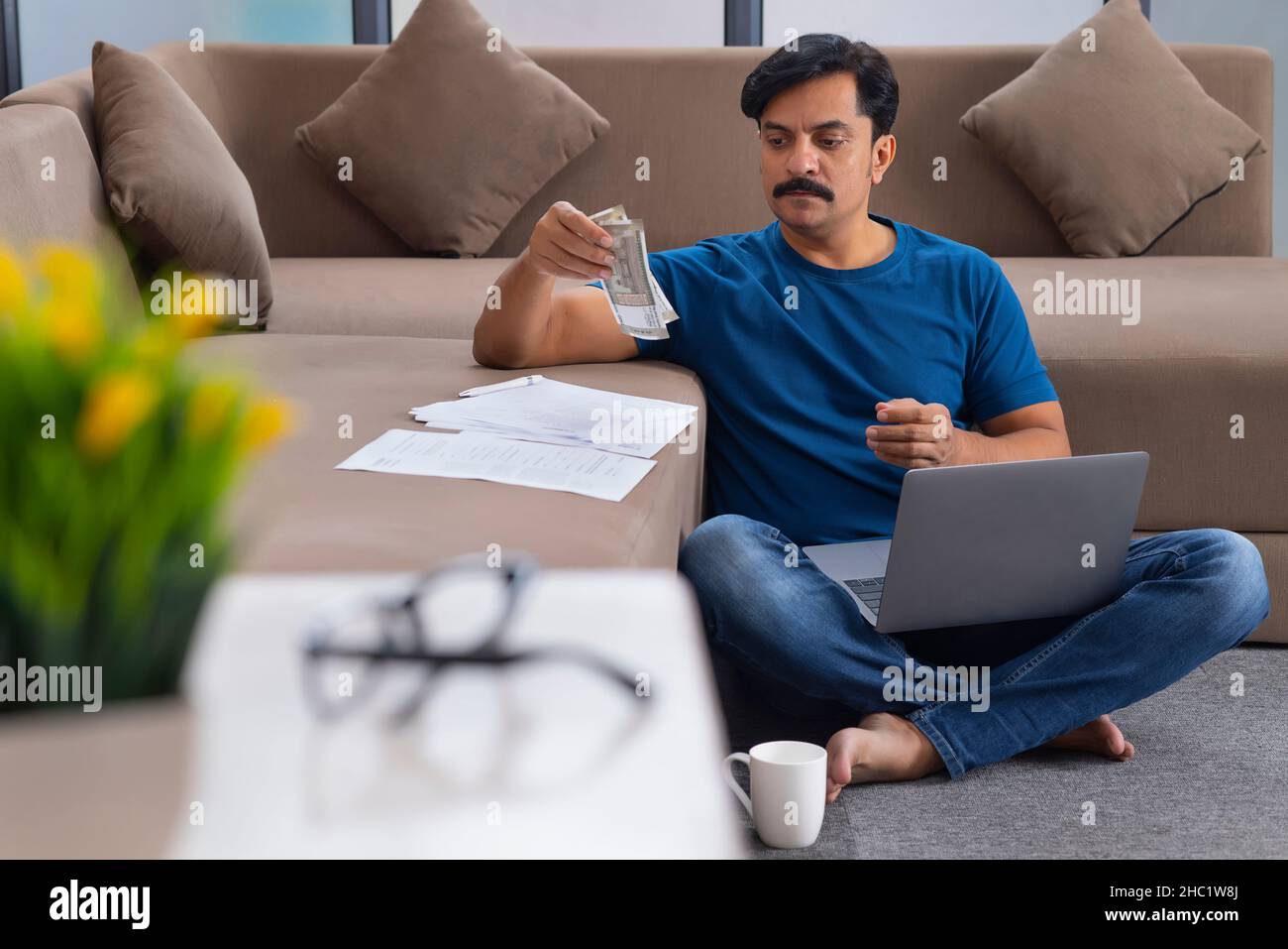 Mann, der indische Währung berechnet, während er auf dem Boden mit einem Laptop auf dem Schoß sitzt Stockfoto