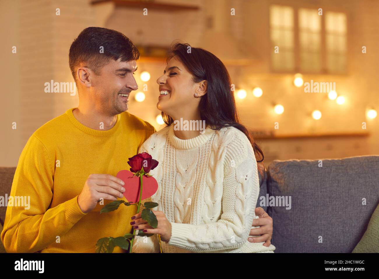 Der Ehemann umarmt seine geliebte Frau und gibt ihr eine Rose und eine Valentinskarte, während er auf der Couch sitzt. Stockfoto