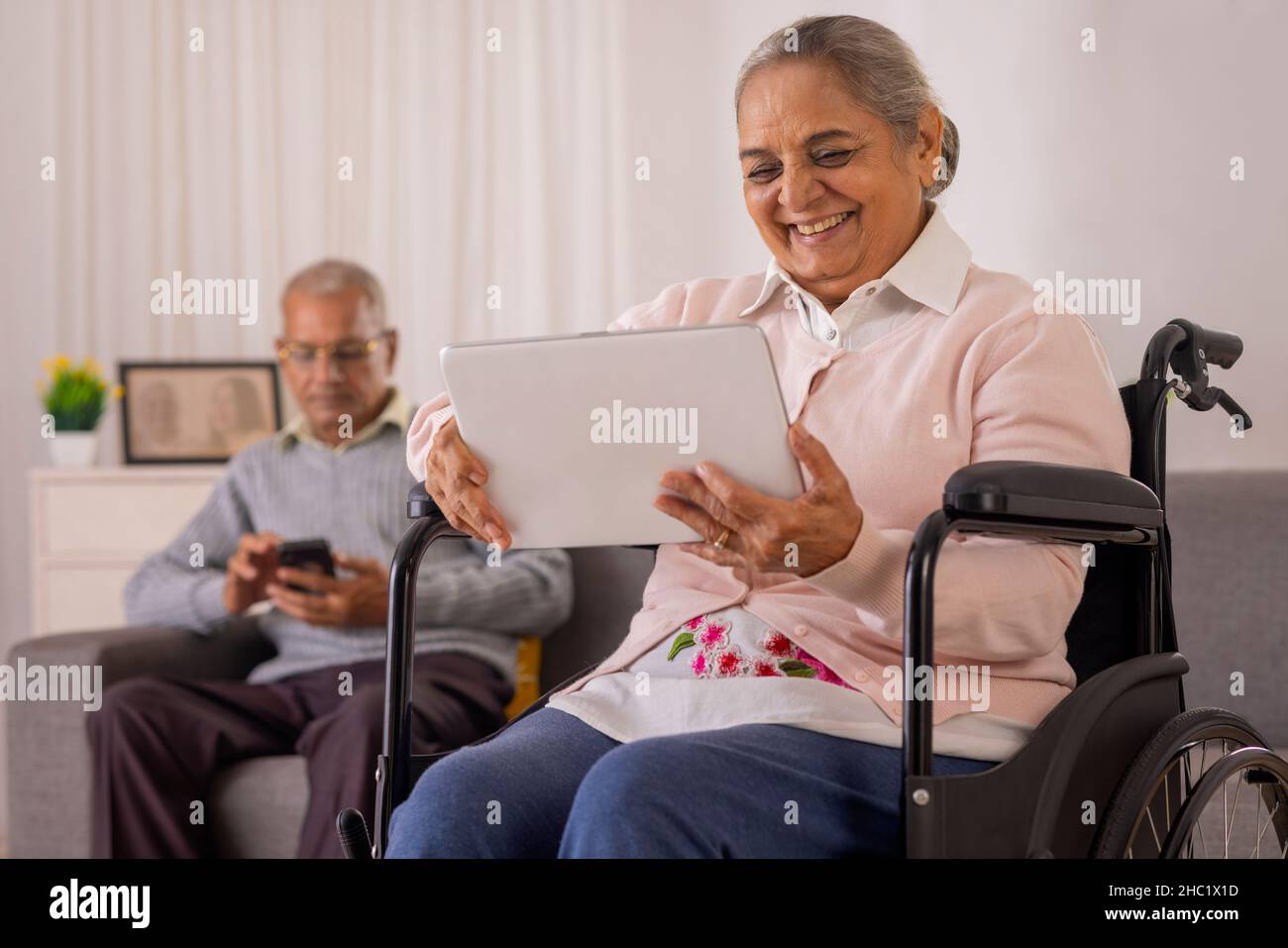 Alte Frau mit digitalem Tablet auf dem Rollstuhl und alter Mann mit Smartphone auf dem Sofa Stockfoto