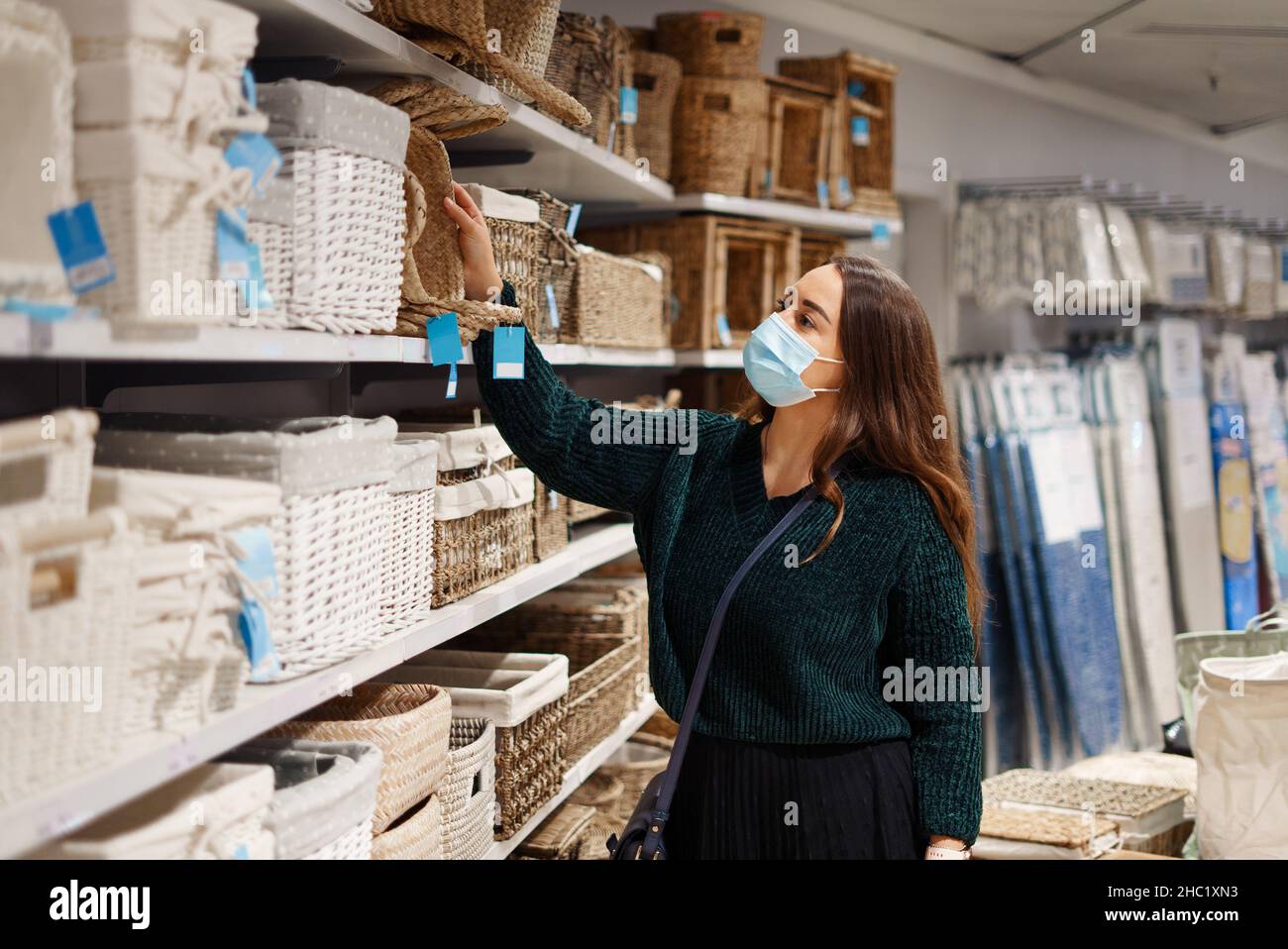 Junge Dame in Gesichtsmaske wählt Weidenkörbe in der Haushaltswarenabteilung in der Store Mall, prüft Preise und Qualität, genießt ruhig ruhig Einkaufen allein während covid19 Pandemie Stockfoto