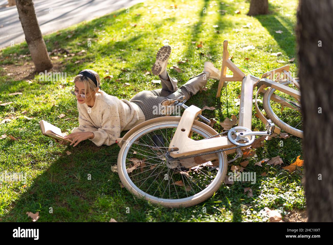 High-Angle-Ganzkörper der Frau in stilvollen Sonnenbrillen liegen in der Nähe von Holz Fahrrad und interessante Geschichte auf grasbewachsenen Lichtung im Park zu lesen Stockfoto