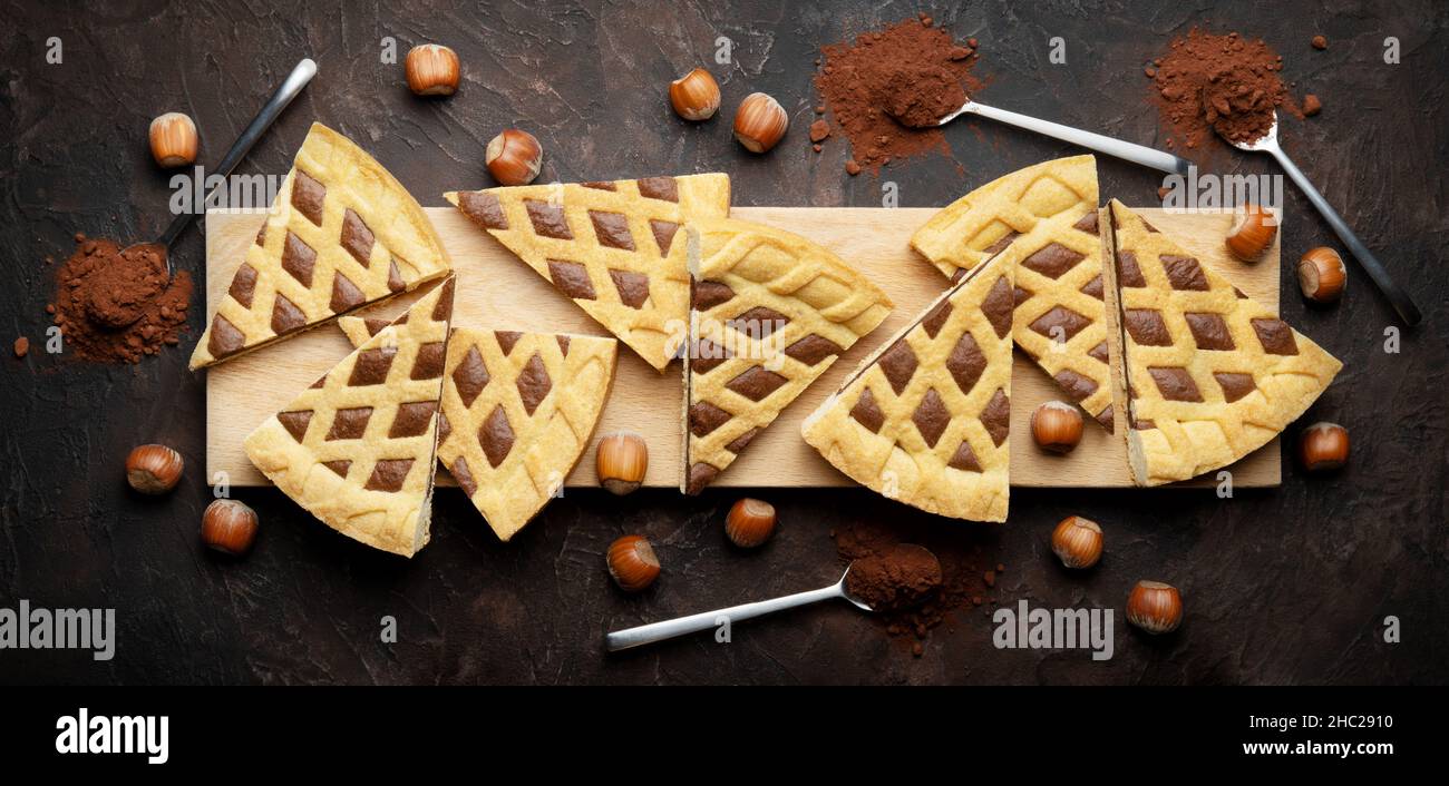 Crostata, acht Scheiben Haselnuss- und Kakaotarte auf Holzschneidebrett mit Löffeln und Kakaopulver. Draufsicht auf braunem Gipshintergrund, flaches Lay Stockfoto