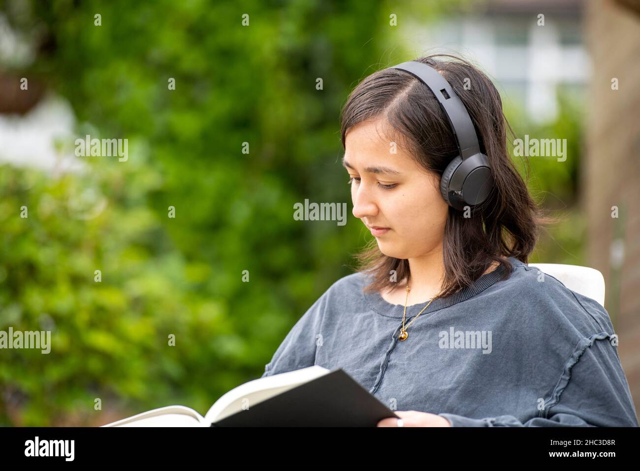 Junge Frau mit Kopfhörern liest Buch im Garten Stockfoto