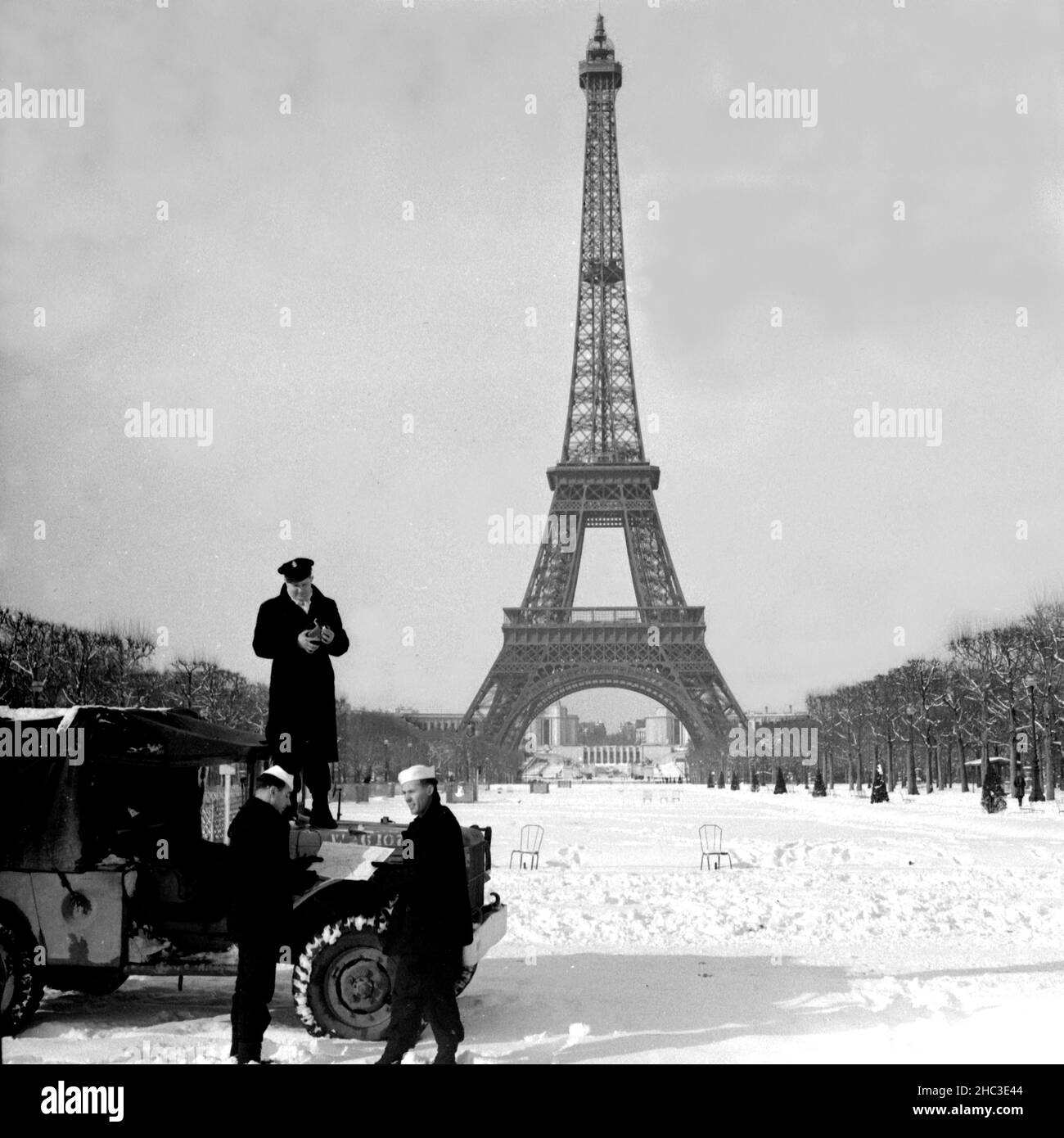 US Navy Männer und Jeep im Schnee im Champs de Mars mit dem Pariser Eiffelturm, 1945. Im vielleicht ersten schweren Schnee nach der Befreiung haben drei amerikanische Marinemänner ihren Jeep mit dem Eiffelturm im Hintergrund auf die Champs de Mars gezogen. Keine anderen Menschen sind in Sicht. Der Turm ist von unten nach oben abgebildet und der Palais de Chaillot ist zwischen den Beinen zu sehen. Ein US-Chef-Kleinoffizier in Peacoat steht mit einer Filmkamera auf der Motorhaube des Jeeps. Zwei Seeleute in Marinekappen und Friedensmützen halten eine Karte von Paris. Stockfoto