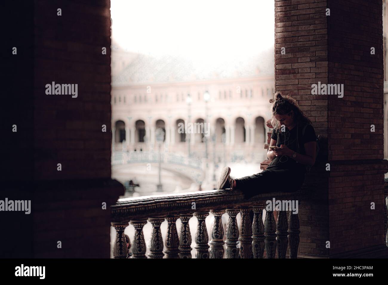 Junge Frau, die auf einem Steinzaun des Gebäudes sitzt und am Telefon tippt Stockfoto