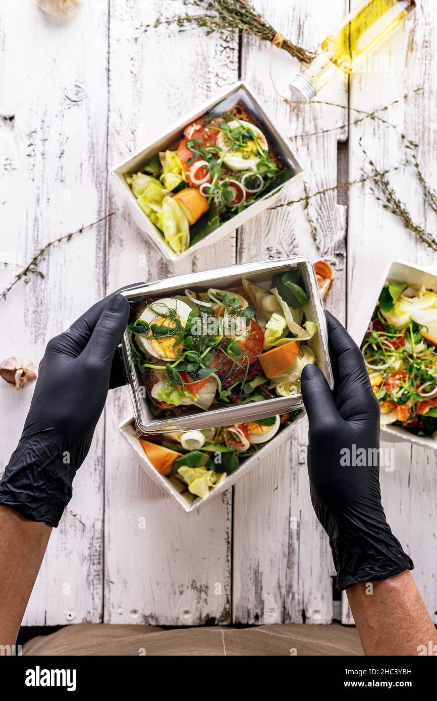 Hände in schwarzen Handschuhen packen ein Business Lunch in einen Karton. Konzept Food Delivery Service. Restaurant zum Mitnehmen. Leckeres Essen bei der Arbeit Stockfoto