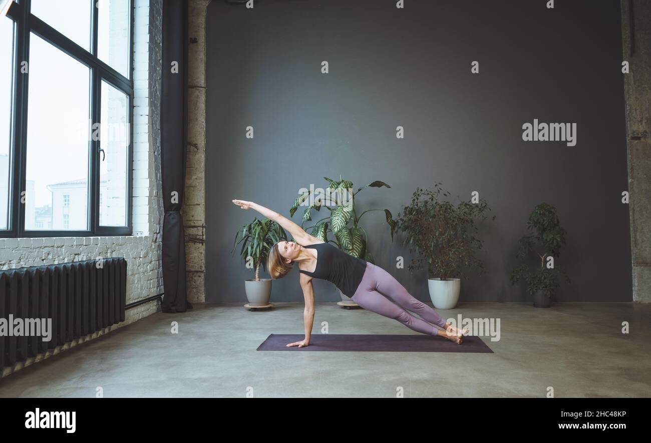 Lonely Woman in Sportswear macht Side Plank in einem geräumigen Yoga Studio. Die Sportliche Frau Praktiziert Hatha Yoga. Volle Länge. Grauer Hintergrund. Hochwertige Fotos Stockfoto