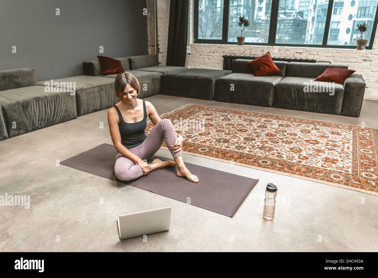 Die sportliche Frau sitzt auf einer Sportmatte und hat während der Quarantänezeit einen Online-Video-Chat mit ihrem Fitness Coach auf dem Laptop. Home Workouts. Stadthintergrund. Nahaufnahme. Hochwertige Fotos Stockfoto