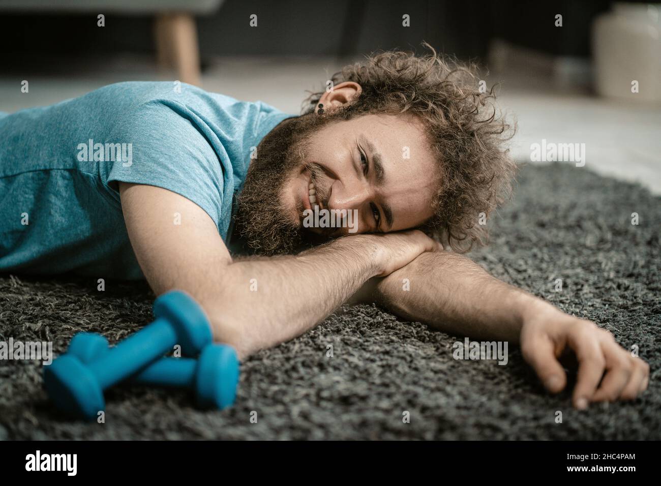 Der Mann liegt entspannt auf dem Teppich neben seinen Hanteln. Der lockige Kerl ist zu entspannt, um Sport zu machen. Nahaufnahme Porträt . Hochwertige Fotos Stockfoto