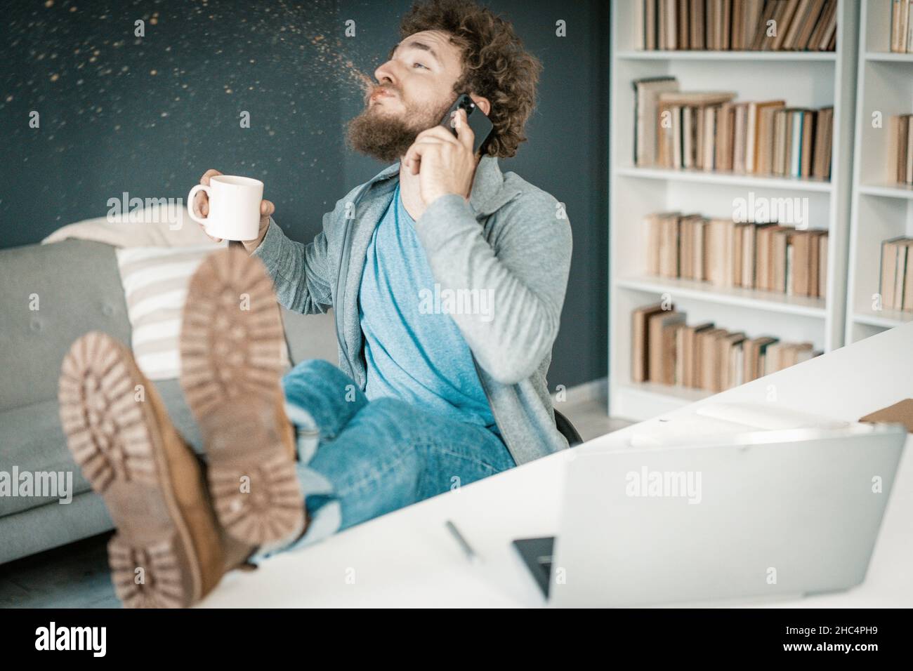 Überraschender Mensch hört die Nachrichten auf seinem Smartphone und sput Kaffee aus. Der Mann hat Beine auf dem Tisch und einen Becher Kaffee in der Hand. Nahaufnahme. Bücherregale Hintergrund. Hochwertige Fotos Stockfoto