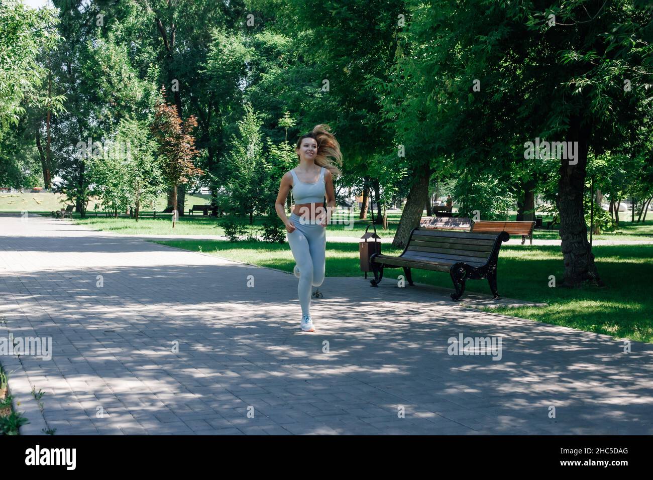 Laufen schnell lächelnd junge sportliche Frau wärmen sich auf, Stretching in grau Sportkleidung im Park an sonnigen Sommertag Stockfoto